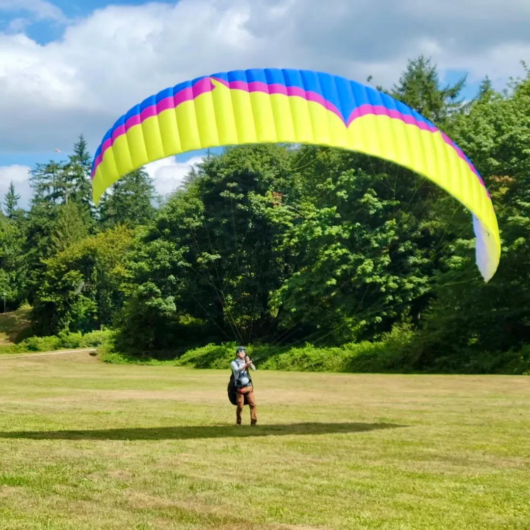 A pilot standing in a grass field under an inflated paraglider wing, forest and clouds behind.